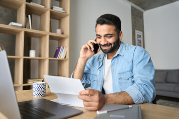Young happy smiling adult indian Hispanic businessman talking on mobile cellphone arranging meeting with employee reading paper CV sitting at desk at home office. Remote recruitment work concept.