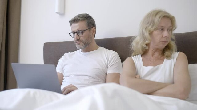 Blond wife lying in bed and trying to talk to busy husband working on laptop