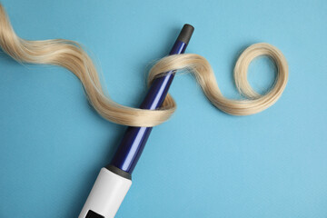 Curling iron with blonde hair lock on light blue background, flat lay