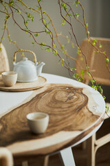 Interior design of stylish dining room interior with family wooden and epoxy table, rattan chairs, flowers in vase and teapot with cups.  Details. Template.