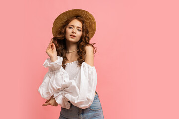 Fashion studio photo of elegant red haired  girl in straw hat and summer outfit posing on pink background.