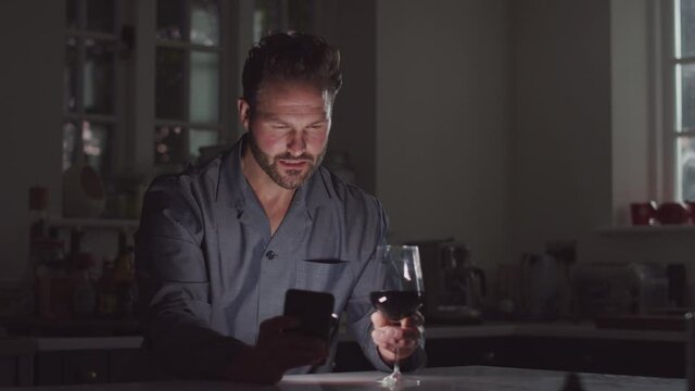 Man wearing pyjamas drinking wine and using mobile phone alone at home at night looking unhappy - shot in slow motion