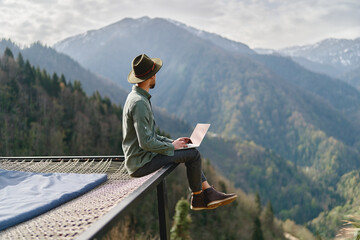 Young man freelancer traveler wearing hat anywhere working online using laptop and enjoying...