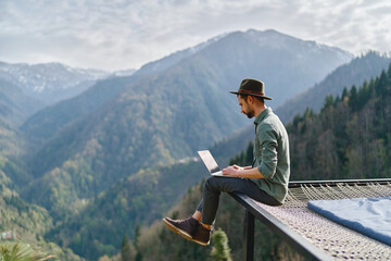 Young man freelancer traveler wearing hat anywhere working online using laptop and enjoying...