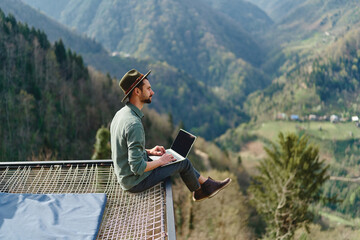 Young man freelancer traveler wearing hat anywhere working online using laptop and enjoying mountains view