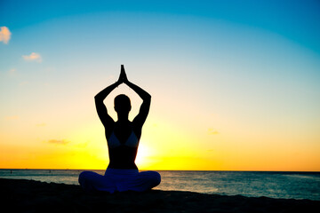 Woman meditating, doing yoga, at the beach, sitting by the seashore, dressed in a white outfit at sunset