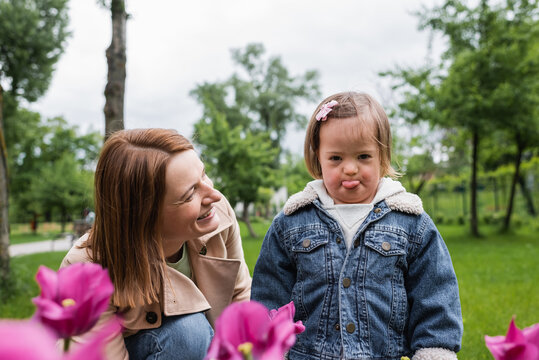happy mother smiling and looking at disabled daughter sticking out tongue