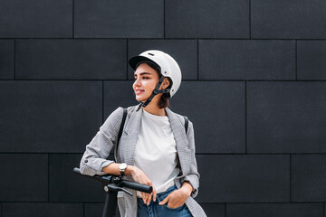 Young woman wearing a scooter helmet standing at a black wall. Smiling female in casual clothes...
