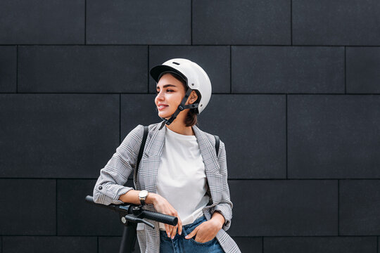 Young woman wearing a scooter helmet standing at a black wall. Smiling female in casual clothes leaning electric scooter looking away.