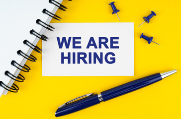 On a yellow background lies a notebook, a pen and a business card with the inscription - WE ARE HIRING