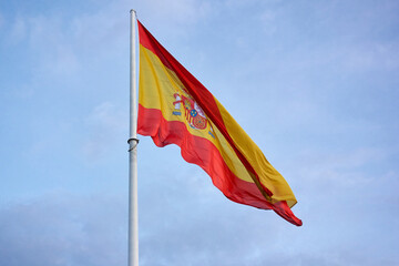 Giant flag of spain hoisted on its mast, on a cloudy day, with blue sky and a lot of wind          ...