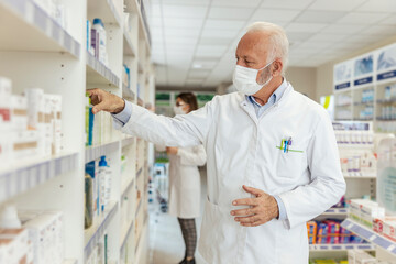 Buying and selling medicines, doctor's advice and help to find medicines on the shelf. Close up of a man with gray hair in a white uniform and a protective face mask sorting drugs at the pharmacy