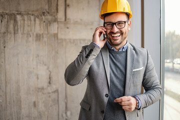 Smiling businessman with helmet on head standing next to a window in building in construction...