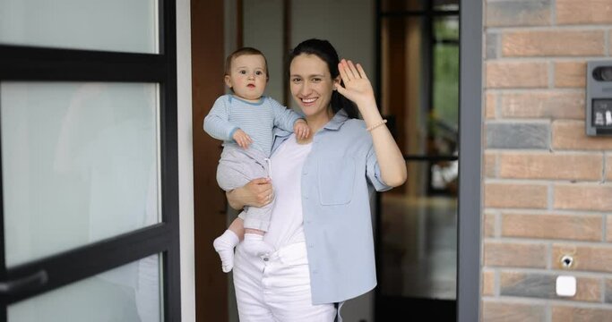 Young Mother With A Baby Waving Hands And Saying Goodbye At Camera While Standing At The Doorway Of The House. Concept Of Farewell With A Family