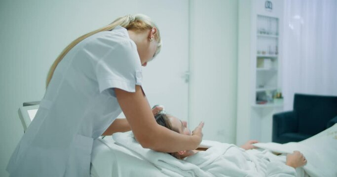 A female cosmetologist is doing a non-injection mesotherapy procedure to face a female client who is laying in the cosmetology bed by using the Dermadrop TDA device.