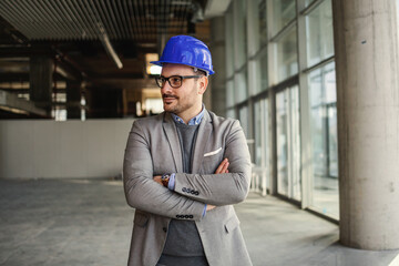 Smiling architect standing in building in construction process with arms crossed.