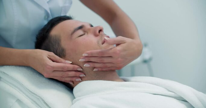 Close-up a male client is laying in the chair while a female cosmetologist is doing a massage of his forehead, temple, cheeks, brow.