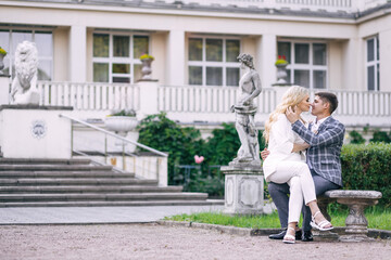 A tender beautiful newlywed couple sits on a bench against the backdrop of greenery and a castle.