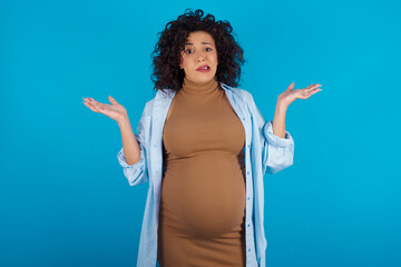 Clueless young Arab pregnant woman wearing dress against blue wall shrugs shoulders with...
