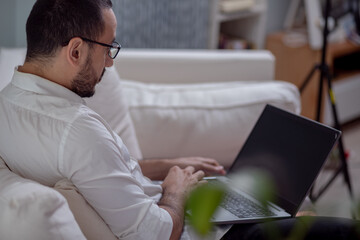 Joven latino trabaja online con su laptop desde la comodidad de su sofá, navegando en internet, y...