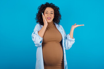 Crazy young Arab pregnant woman wearing dress against blue wall advising discount prices hold open palm new product