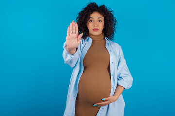 young Arab pregnant woman wearing dress against blue wall doing stop sing with palm of the hand....