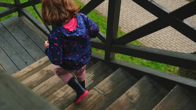 Caucasian Girl Running Down Cabin External Wooden Stairs Wearing Rain Jacket and Gum Welligtons Shoes