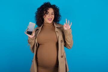 young Arab pregnant woman wearing dress against blue wall holding in hands cell showing ok-sign