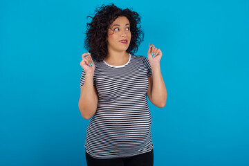 young Arab pregnant woman wearing stripped T-shirt  against blue wall clenches fists and awaits for...