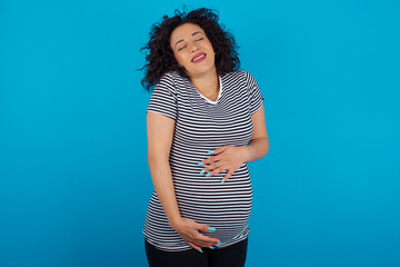 Positive young Arab pregnant woman wearing stripped T-shirt  against blue wall with overjoyed expression closes eyes and laughs shows white perfect teeth