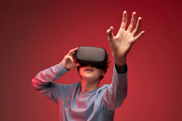 Portrait of young teen boy in VR helmet looks away at copy space, isolated in studio, guy holds out hand forward. Gamer playing VR games on red background, VR gaming. focus on hands