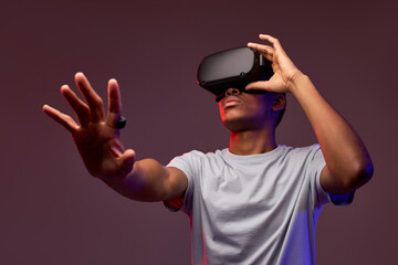 Portrait of young african teen boy in VR helmet looks away at copy space, isolated in studio, guy holds out hand forward. Gamer playing VR games on colourful background, VR gaming.