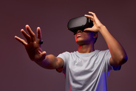 Portrait of young african teen boy in VR helmet looks away at copy space, isolated in studio, guy holds out hand forward. Gamer playing VR games on colourful background, VR gaming.
