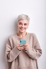 Adorable Female With Blonde Short Hair Chatting With Someone On Mobile Phone, Smiling Cutely, Looking At Side Thinking How To Reply, What To Write. Isolated White Studio Background