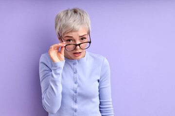 young Woman in glasses looks through them at camera, looks confused, analyzing situation. serious woman expresses doubt and suspicion, feeling sceptical about something