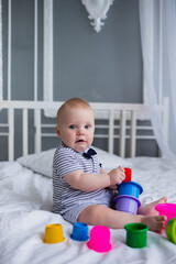 caucasian baby boy with blue eyes in a striped jumpsuit sits on the bed and plays with plastic toys. Early child development
