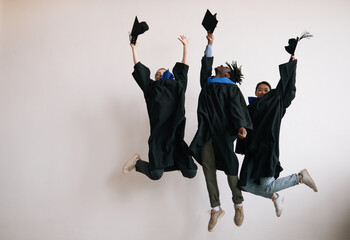 Full length of three happy college graduates wearing robes and jumping against white background,...