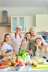 Positive caucasian women children guhhing each other in bright cozy kitchen, posing at camera, spending ggod holidays together, wearing aprons. copy space