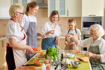 Fototapeta na wymiar Caucasian family preparing delicious tasty pizza in bright modern kitchen, cutting products and adding ingredients, wearing apron.culinary concept. Grandmothers teach young girl to cook
