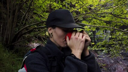 young woman in cap enjoying a drink in a red mug and watching a beautiful waterfall in the forest during a break on her route through the mountains. activities in the mountains. weekend getaway