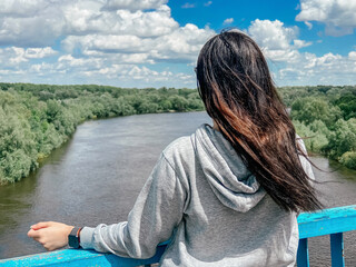 Fototapeta premium A dark-haired сaucasian girl stands with her back in a light gray jacket and looks towards the river and the forest in the city of Chernigov in Ukraine.