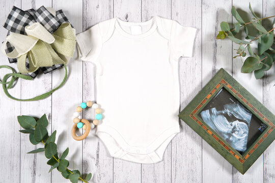 Baby announcement with romper onesie with sonogram flatlay. On-trend farmhouse theme craft product mockup on a white wood background. Negative copy space for your design here.