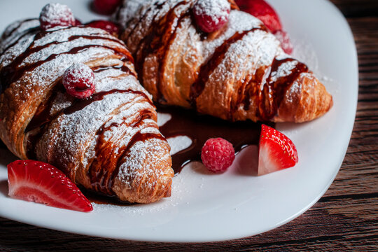 Close up image of a crossaints or croissants on a white plate with strawberries, raspberries and sugar glass or candy glass with chocolate syrup on top 