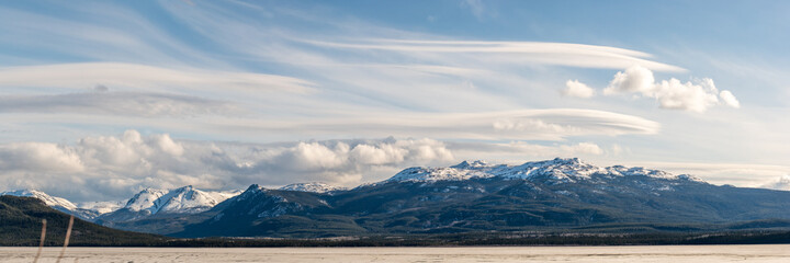 Panorama of snow capped mountains in distance over a frozen lake and amazing, wispy clouds during spring time in northern Canada. 