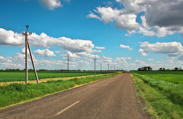 Village Road with power line, green fields and blue sky with clouds