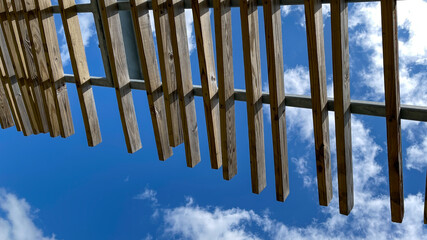 A wooden pergola roof at a park with bright blue skies and white  clouds.