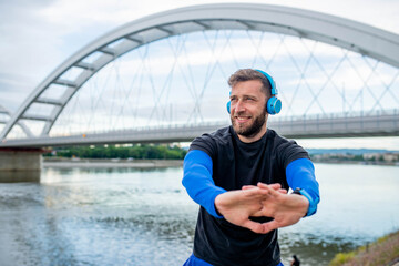 A smiling jogger stretches after running. He listens to music on headphones and in the background...