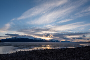 Obraz premium Scenic landscape in northern Canada during spring time with rocks, snow capped mountains in distance in calm lake water on cloudy pink afternoon sunset. 