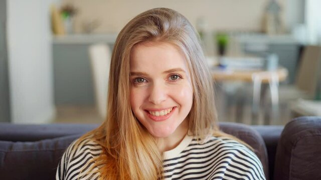 Head and shoulders portrait of beautiful shy young woman with green eyes and in striped sweatshirt looking at camera and smiling timidly sitting on sofa at home
