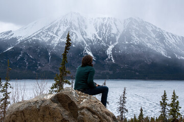 Man sitting on a rock overlooking Yukon Territory landscape during spring time with snow falling on the snow capped peaks in the distance in Canada. 